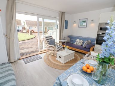 A living room with a sliding glass door opening to a patio and garden with wicker chairs a blue sofa a white coffee table and a dining table with blue flowers and fruit at Little Beach in Seaview