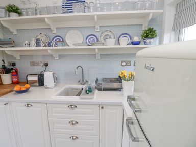 A kitchen counter with a sink a toaster a kettle open shelves with plates mugs and glasses and a refrigerator at Little Beach in Seaview