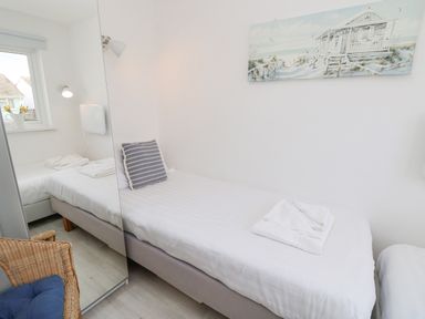 A small bedroom with a single bed white linens a striped pillow and a folded towel on the bed a wicker chair with a blue cushion a bedside lamp a window and a beach painting on the wall at Little Beach in Seaview
