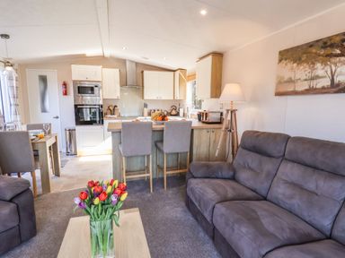 A living room with gray sofas and a wooden coffee table with flowers next to a kitchen with a breakfast bar and stools at Marigold Lodge in Runswick Bay near Staithes