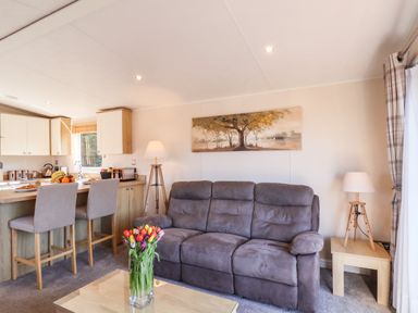A living room with a brown sofa a coffee table with flowers two lamps stools at a kitchen counter and a painting on the wall at Marigold Lodge in Runswick Bay near Staithes