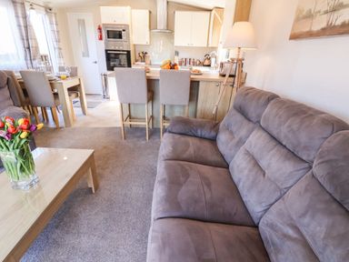 A living room with a sofa and a coffee table with flowers next to a kitchen with a dining table and chairs at Marigold Lodge in Runswick Bay near Staithes