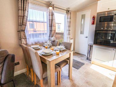 A dining area with a wooden table set with plates glasses and orange juice four upholstered chairs window with curtains and a kitchen with built in microwave and oven at Marigold Lodge in Runswick Bay near Staithes