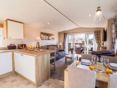 A kitchen and dining area with a wooden table set for breakfast a living room with sofas and a coffee table and sliding glass doors leading to an outdoor seating area at Marigold Lodge in Runswick Bay near Staithes