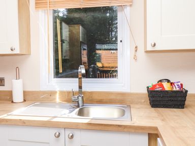 A kitchen countertop with a sink and faucet below a window with a wooden blind and a basket of snacks on the side at Marigold Lodge in Runswick Bay near Staithes
