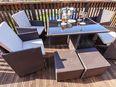 An outdoor dining area with a glass table holding drinks and a bowl of snacks surrounded by wicker chairs on a wooden deck at Marigold Lodge in Runswick Bay near Staithes