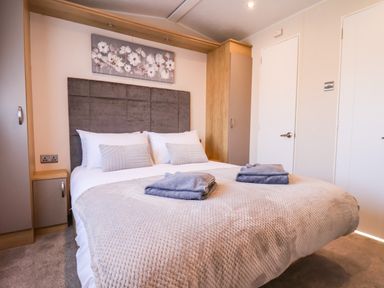 A bedroom with a double bed, two folded towels on the bed, wardrobe cabinets, and a floral painting above the headboard at Marigold Lodge in Runswick Bay near Staithes
