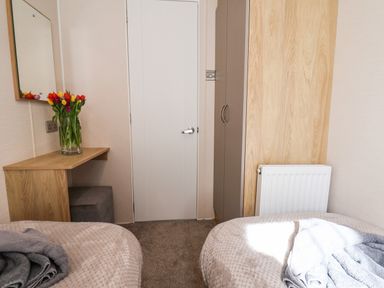 A bedroom with two beds a wooden dressing table with a vase of flowers a wall mirror a white door and a wardrobe at Marigold Lodge in Runswick Bay near Staithes