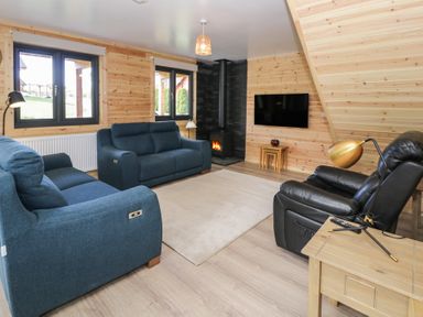 A living room with blue sofas a black leather chair a wood burning stove and a wall mounted tv at Sunset Lodge 5 in Llanbedr