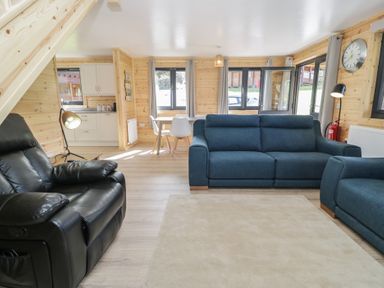 A living room with blue sofas a black recliner a dining table with white chairs and a kitchen area at Sunset Lodge 5 in Llanbedr