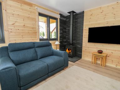 A living room with a blue sofa a wood burning stove a flat screen tv on the wall nested wooden tables and windows at Sunset Lodge 5 in Llanbedr