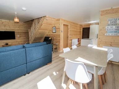 A dining area with a wooden table and six white chairs next to a living area with blue sofas and a black chair at Sunset Lodge 5 in Llanbedr