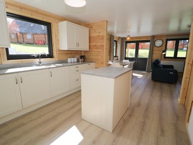 A kitchen area with countertops a sink and white cabinets leading to a dining area with a table and chairs and a living area with a blue sofa and clock on the wall at Sunset Lodge 5 in Llanbedr