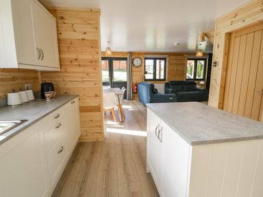 A kitchen with white cabinets and gray countertops leading to a dining area with white chairs and a living room with two blue sofas at Sunset Lodge 5 in Llanbedr