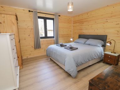 A bedroom with a double bed with gray bedding two bedside lamps a white dresser a wooden chest and a window with gray curtains at Sunset Lodge 5 in Llanbedr