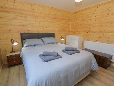A bedroom with a double bed with grey bedding and towels on top two bedside tables with lamps a white chest of drawers and wood-paneled walls at Sunset Lodge 5 in Llanbedr