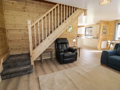 A living room with wooden staircase a black armchair and kitchen in the background at Sunset Lodge 5 Llanbedr