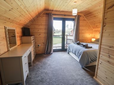 A bedroom with wooden walls a bed with towels a dresser a TV a mirror and glass doors leading outside at Sunset Lodge 5 in Llanbedr