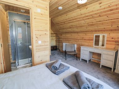 A bedroom with wooden walls a bed with towels a grey chair a dressing table with a mirror and a bathroom with a shower at Sunset Lodge 5 in Llanbedr