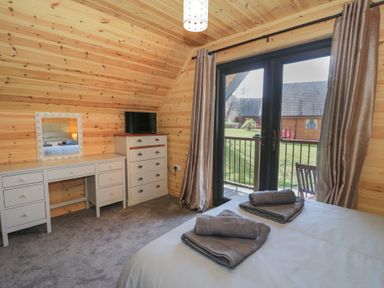 A bedroom with a bed, dresser, desk with mirror and TV next to glass doors leading outside at Sunset Lodge 5 in Llanbedr