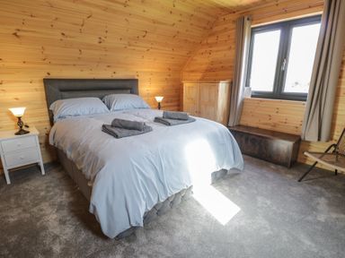 A bedroom with a double bed with folded towels bedside tables lamps a wooden cabinet a bench and a window at Sunset Lodge 5 in Llanbedr