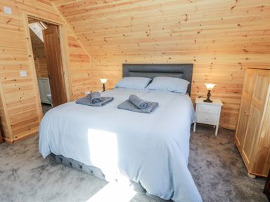 A bedroom with a double bed with gray pillows and towels beside a wooden wardrobe and bedside tables with lamps at Sunset Lodge 5 in Llanbedr