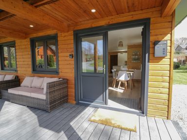 An outdoor porch with a wicker sofa and cushions next to a door opening into a dining area with a table and chairs at Sunset Lodge 5 in Llanbedr