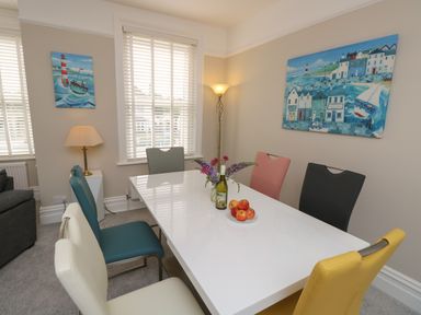 A dining room with a white table surrounded by six multicolored chairs and two paintings on the walls at Chine Heights in Bournemouth