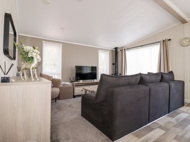 A living room with a large dark sofa a television two windows and a sideboard with decorations at 63 The Maples in Hewas Water near Sticker