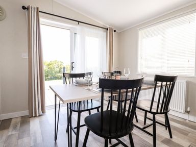 A dining room with a wooden table set with plates and glasses and black chairs near large windows at 63 The Maples in Hewas Water near Sticker