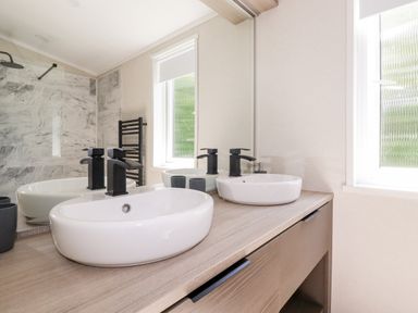 A bathroom with two white oval sinks on a wooden countertop a window and a marble shower wall at 63 The Maples in Hewas Water near Sticker