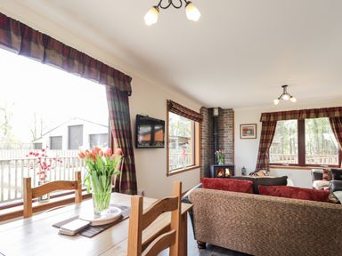 A living room with a dining table with flowers and a wood stove next to couches and windows with plaid curtains at Lovat Highland Bothy in Beauly