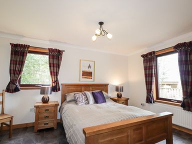 A bedroom with a wooden bed and two side tables with lamps and two windows with plaid curtains at Lovat Highland Bothy in Beauly