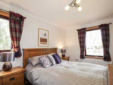 A bedroom with a wooden bed and bedside tables next to windows with plaid curtains at Lovat Highland Bothy in Beauly