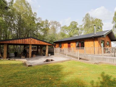A wooden cabin with a fenced porch and a covered outdoor seating area surrounded by trees at Lovat Highland Bothy in Beauly