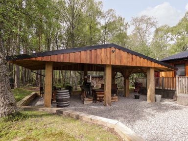 An outdoor wooden pavilion with picnic tables and a barrel in a forested area at Lovat Highland Bothy in Beauly