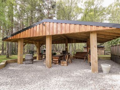 A wooden outdoor shelter with picnic table and benches in a forested area at Lovat Highland Bothy in Beauly