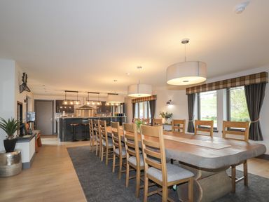 A dining room with a long wooden table surrounded by chairs and a kitchen area with bar stools at Ben Luna Lodge in Drumnadrochit Loch Ness