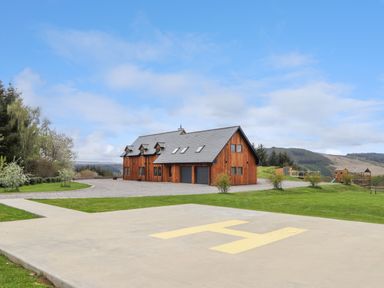 A wooden house with a gray roof next to a helipad and green lawn at Ben Luna Lodge in Drumnadrochit Loch Ness