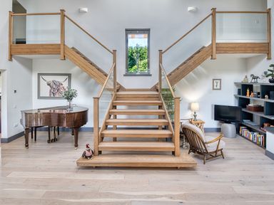 An interior with a wooden staircase leading up with glass railings a piano on the left and a reading area with a chair a lamp and a bookshelf on the right at Ben Luna Lodge Drumnadrochit Loch Ness