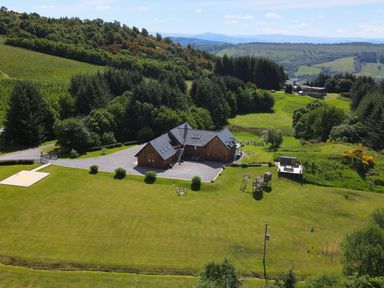 A large wooden house with a gravel driveway and surrounding green lawn with playground equipment and a helipad on a hillside at Ben Luna Lodge in Drumnadrochit Loch Ness