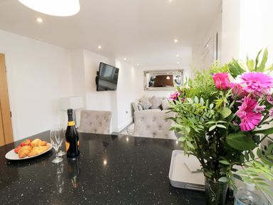A kitchen counter with a bottle of wine glasses and croissants with a living room and a sofa visible beyond at Tower Apartment in Whitby