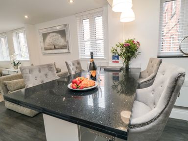 A dining area with a black table top surrounded by upholstered chairs a plate with croissants and strawberries a bottle of rose wine two wine glasses flowers in a vase and windows with blinds at Tower Apartment in Whitby
