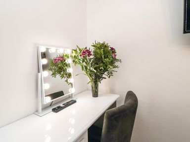 A white desk with a lighted mirror a remote and a vase with flowers and a dark chair at Tower Apartment in Whitby