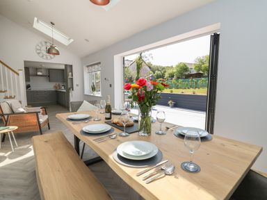 A dining area with a wooden table set for six with flowers and a bottle with an open plan kitchen in the background at Rhiwmynach in Pontarfynach near Aberystwyth