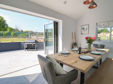 A dining area with a wooden table set for six people next to open doors leading to an outdoor patio with a barbecue grill at Rhiwmynach in Pontarfynach near Aberystwyth