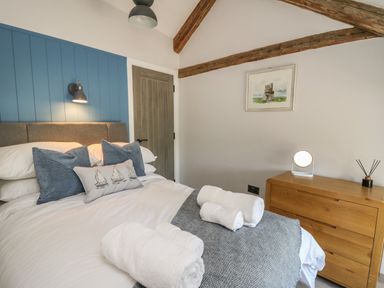 A bedroom with a bed with pillows and rolled towels beside a wooden dresser with a lamp and artwork on the wall at Rhiwmynach in Pontarfynach near Aberystwyth