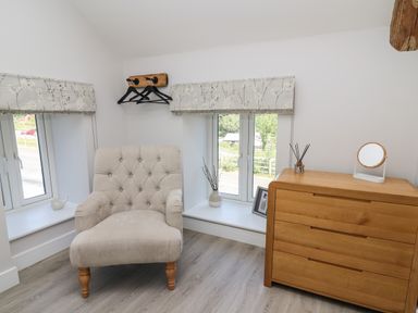 A corner room with a beige armchair next to two windows and a wooden chest of drawers with a mirror on top at Rhiwmynach in Pontarfynach near Aberystwyth
