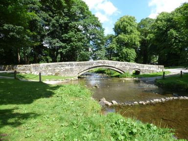 A stone arch bridge over a shallow river with green grass and trees surrounding at The Folly in Linton near Thresfield