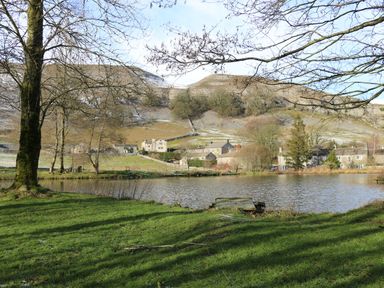 A pond with a wooden dock and grass in front of houses and hills at The Folly in Linton near Thresfield